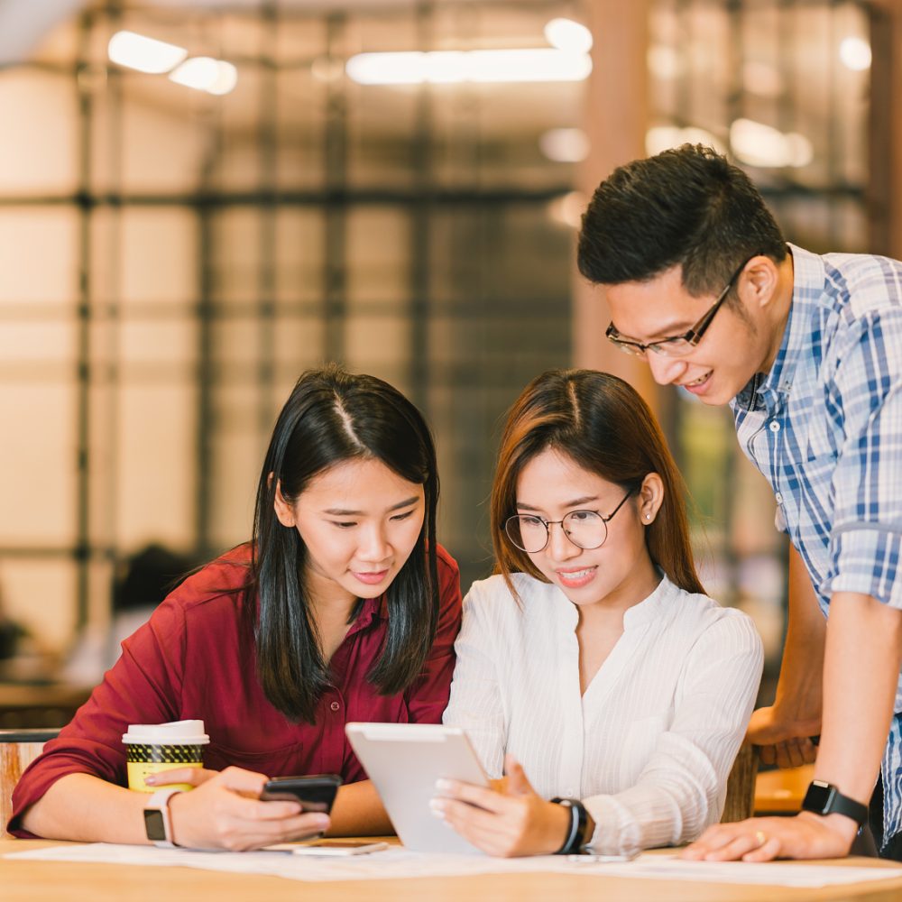 Young Asian college students or coworkers using digital tablet and smartphone together at coffee shop, diverse group. Casual business, freelance work at cafe, social meeting, or education concept